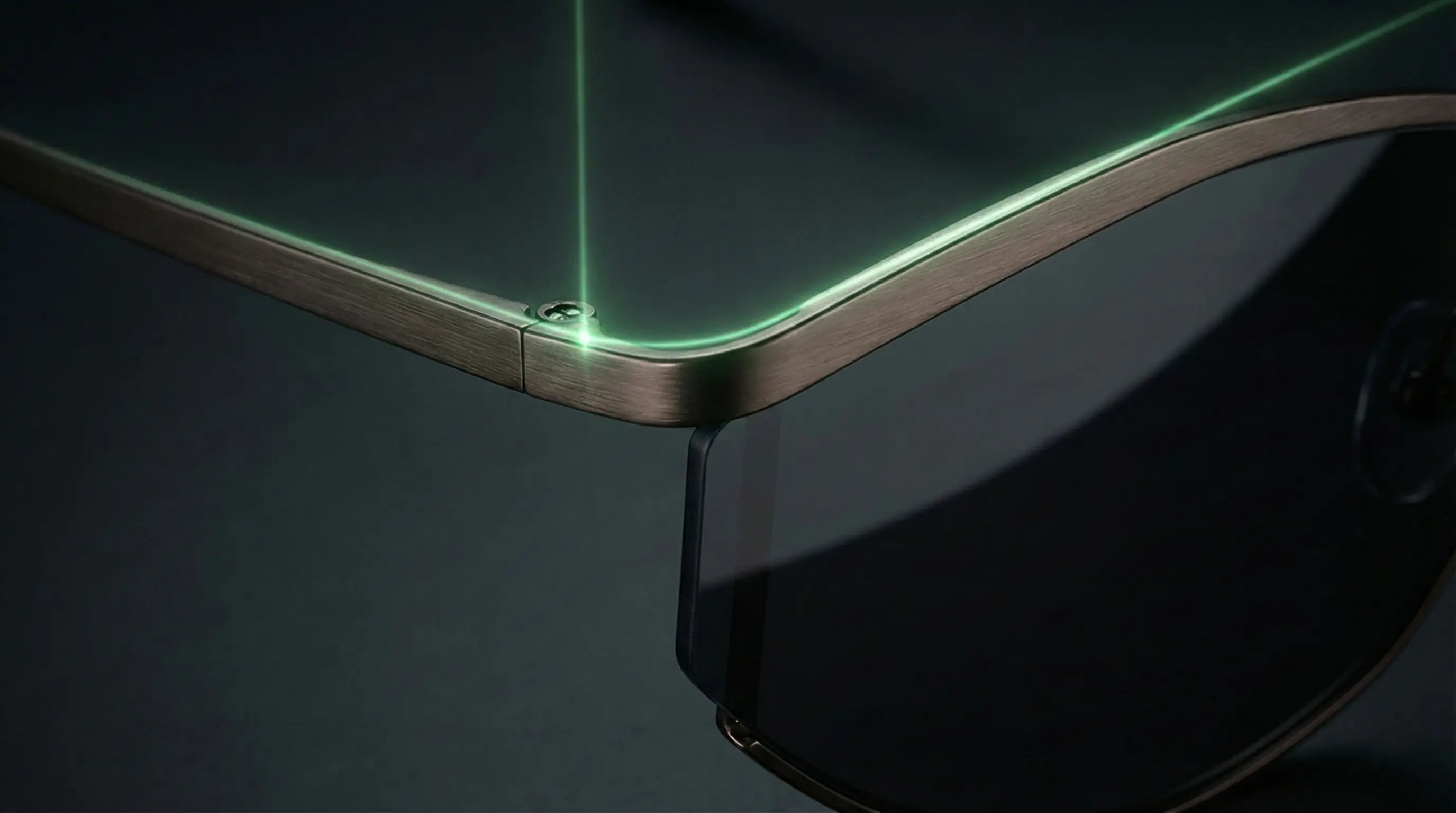 Close-up of a sunglasses frame (brushed metal finish) with a green laser highlighting the hinge detail, set against a dark background to emphasize sleek, technical design.