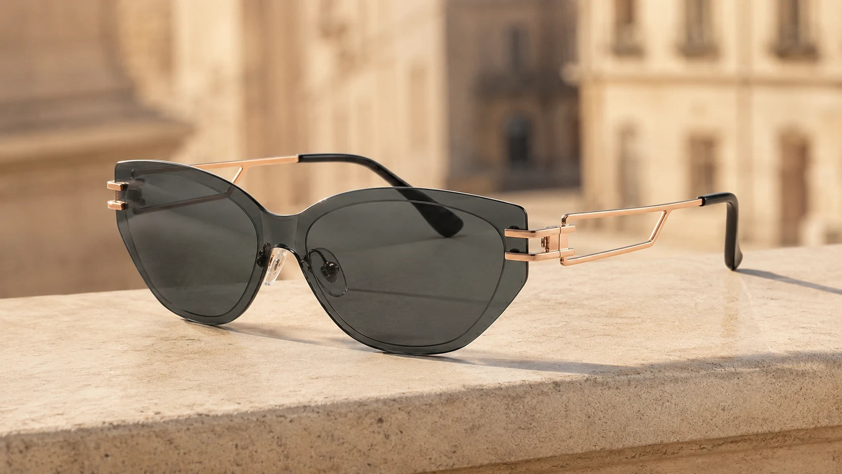 BAPORSSA Edge sunglasses with smoke grey cat-eye lenses and rose gold
geometric temples, resting on a stone ledge with a blurred European
architectural background.