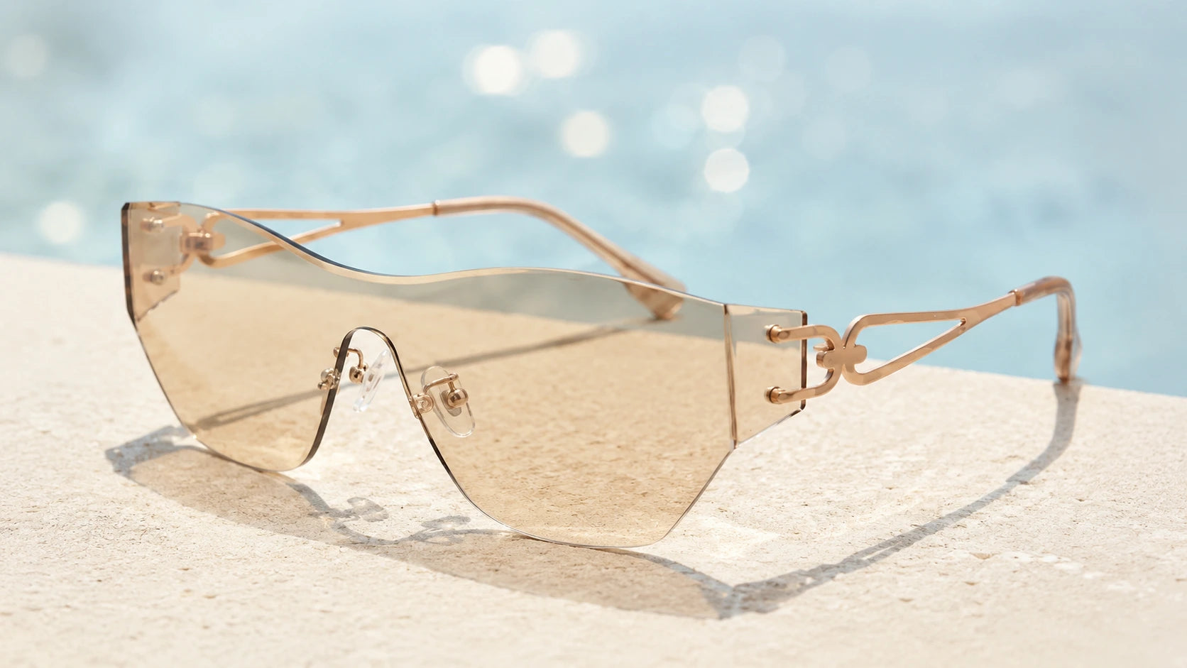 BAPORSSA rimless sunglasses with champagne-tinted lenses and rose gold
decorative temples, resting on a stone edge with a blurred swimming pool
background