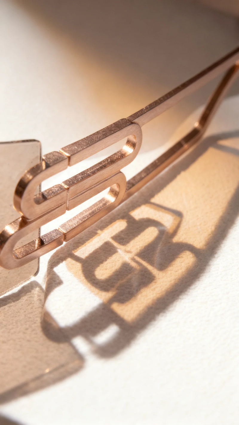 Close-up of a sunglasses frame featuring a rose gold, textured paperclip-style sculptural detail; warm light casts a shadow of the design onto a soft surface, highlighting the modern, artistic craftsmanship.