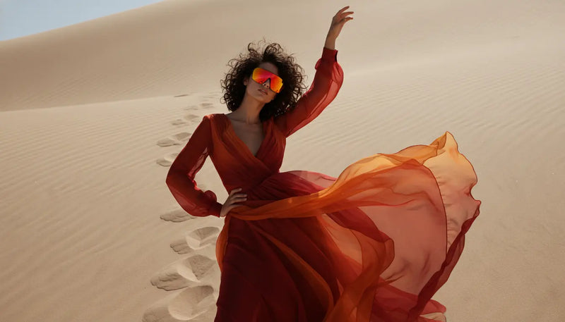 A woman with dark, curly hair wearing a deep red and orange ombre flowing maxi dress and bright red/orange mirrored visor sunglasses, posing dramatically on a sand dune with footprints visible in the background.