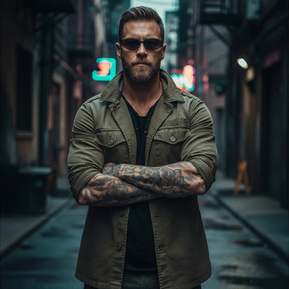 A bearded man with arm tattoos, wearing a green jacket and sunglasses, stands with arms crossed in a neon-lit city alley.