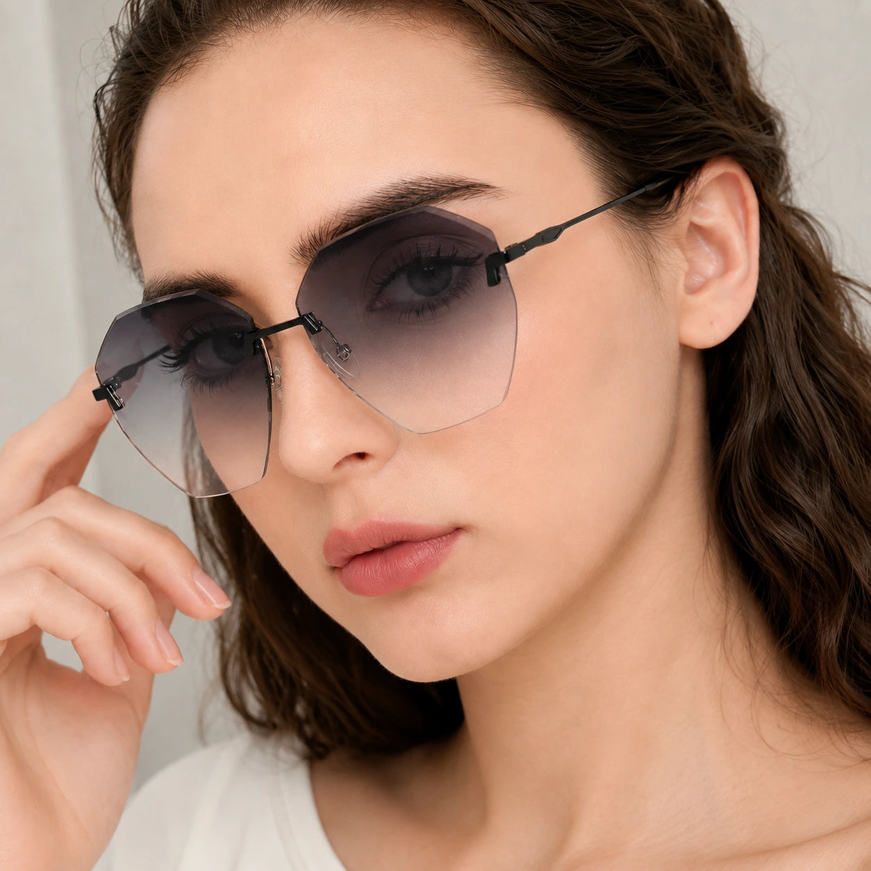 A tight close-up portrait of a woman adjusting her Baporssa Glow rimless sunglasses, featuring black hardware and dark gray gradient lenses.