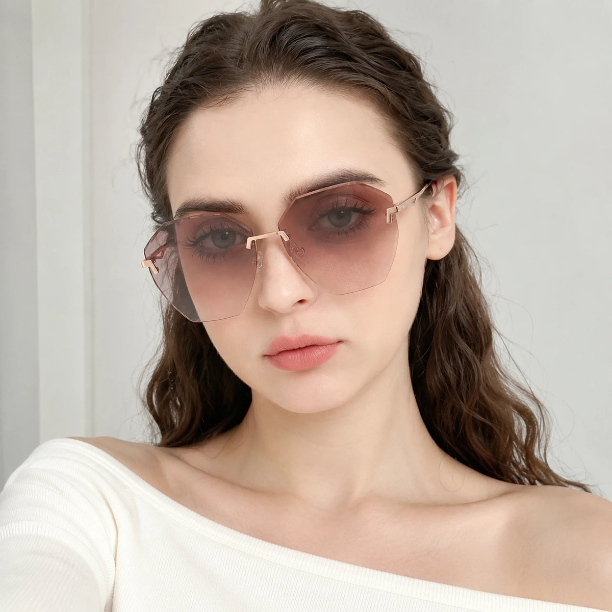 A direct front view portrait of a woman wearing Baporssa Glow rimless sunglasses with pinkish-brown gradient polygonal lenses and an off-the-shoulder white top.
