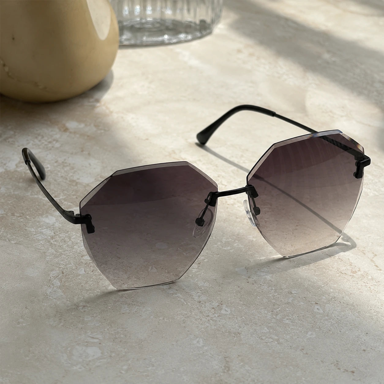 Baporssa Glow rimless polygonal sunglasses with dark gray gradient lenses and black metal hardware, resting on a sunlit beige marble counter with soft shadows.