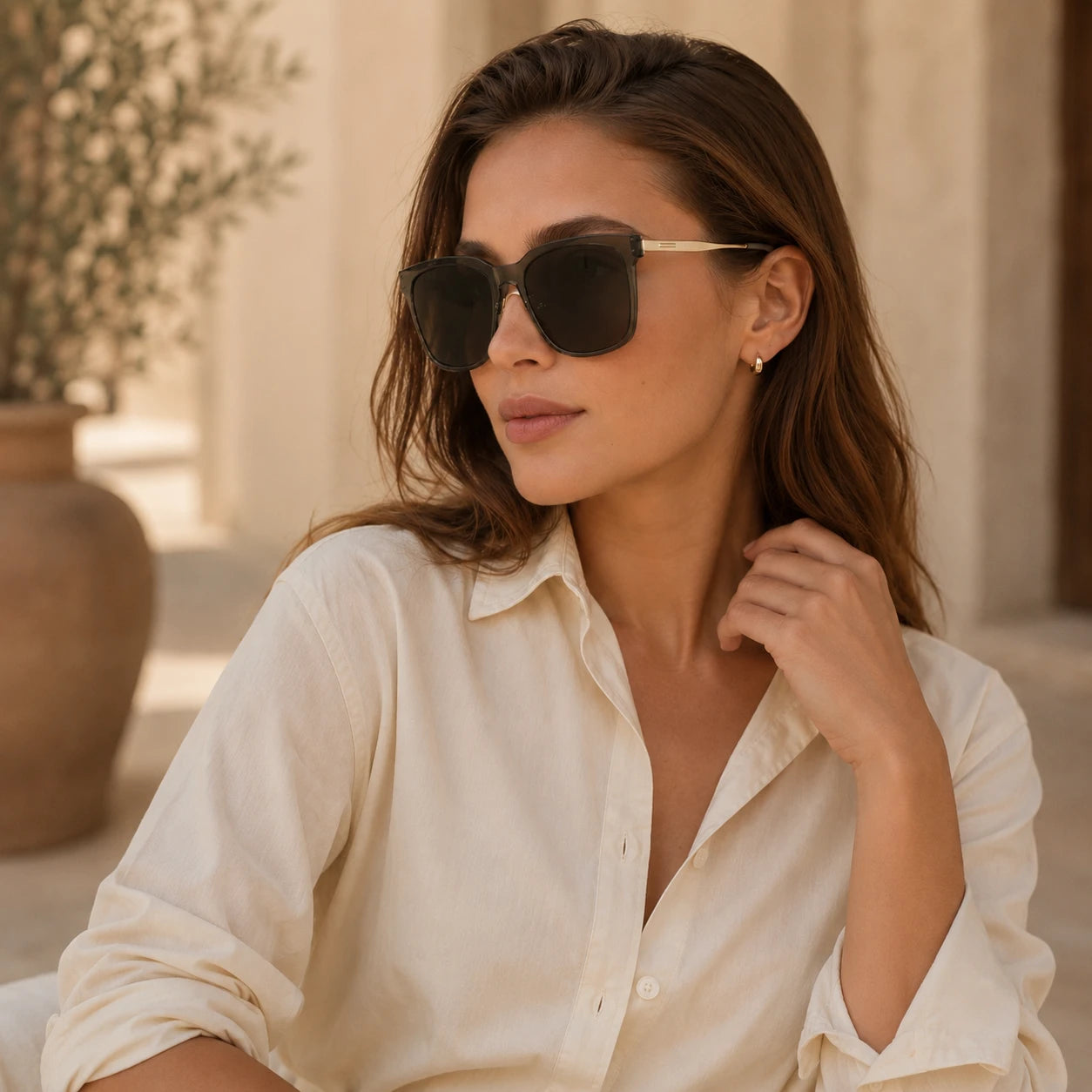 A lifestyle portrait of a woman wearing Baporssa Halo transparent grey sunglasses and a cream button-down shirt, sitting in a sunlit outdoor setting.