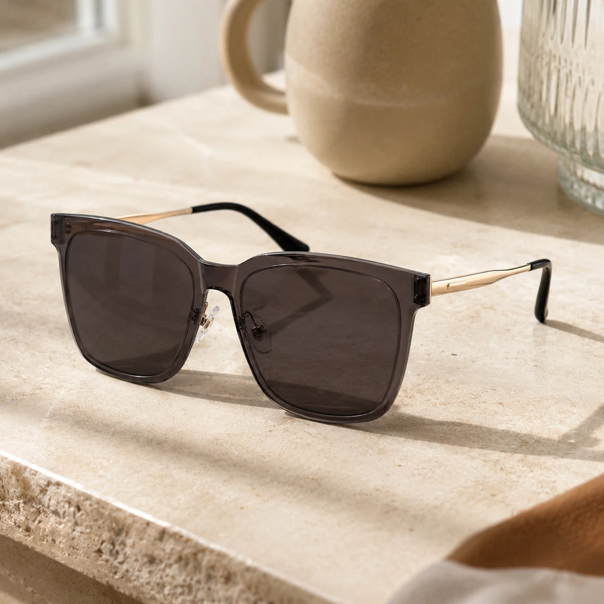 Baporssa Halo square sunglasses with dark lenses and gold temples, displayed on a sunlit beige marble surface next to a ceramic mug.