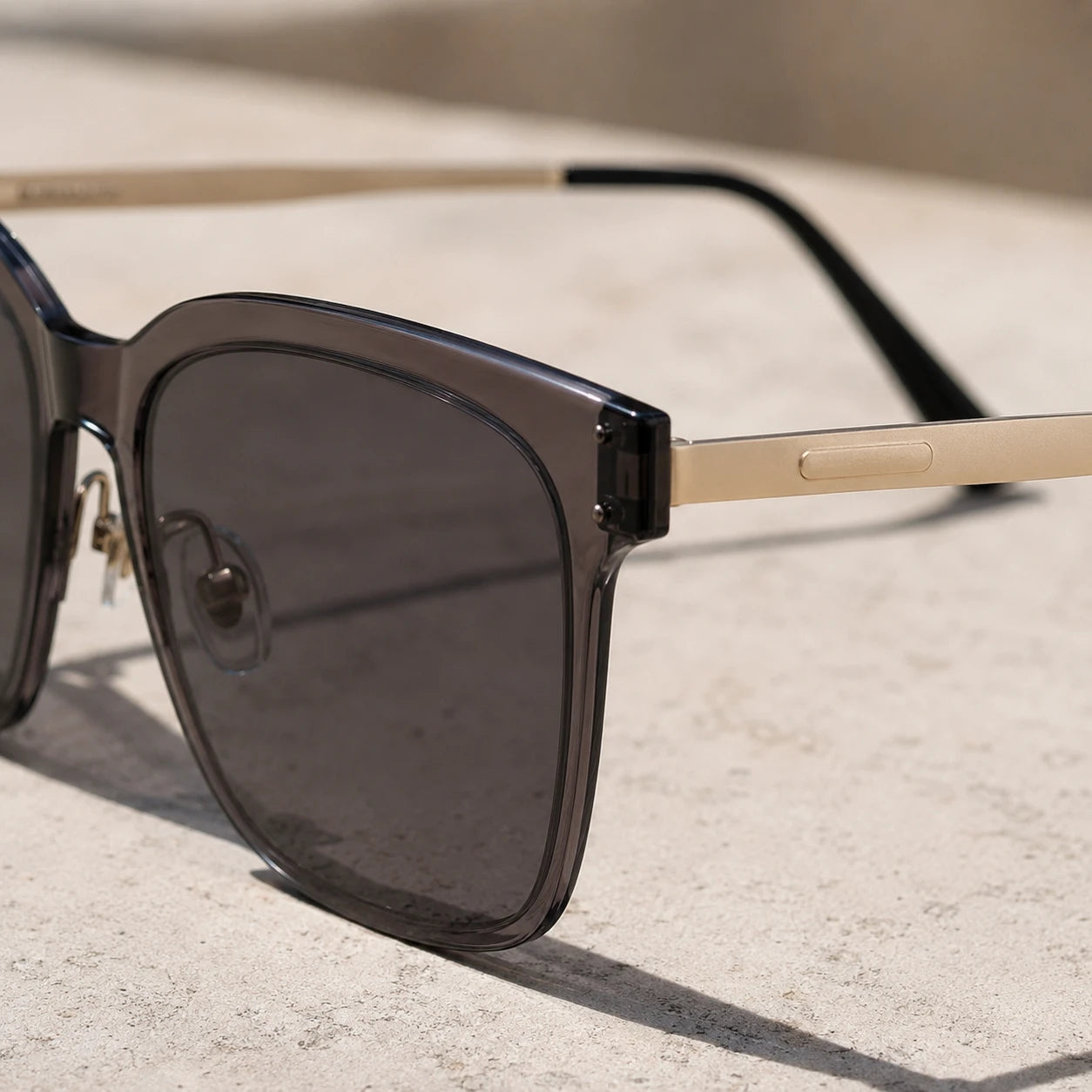 A tight close-up shot focusing on the hinge area of Baporssa Halo square sunglasses, highlighting the translucent grey acetate frame, dark lens, gold metal temple, and the metal nose pad hardware.
