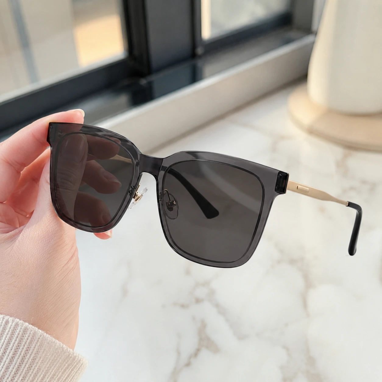 A close-up of a hand holding Baporssa Halo sunglasses, showcasing the translucent grey acetate frame and gold metal hinges.