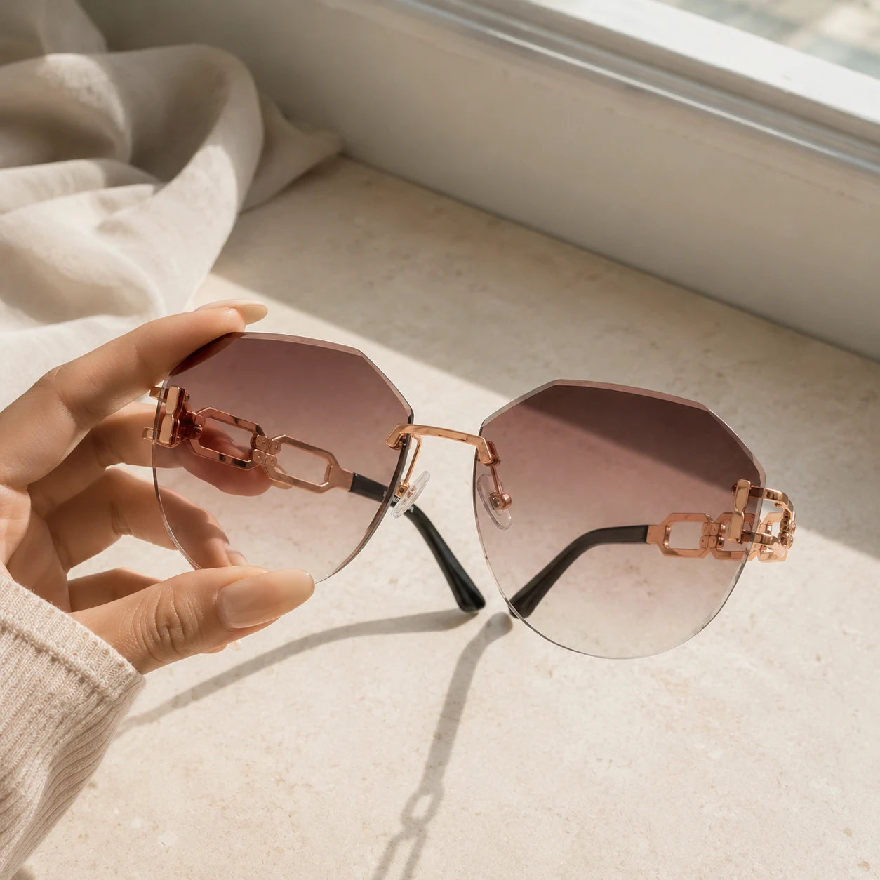 A close-up shot of a hand with manicured nails holding Baporssa rimless sunglasses, showcasing the brown gradient cut-edge lenses and intricate rose gold chain-link arms near a window.