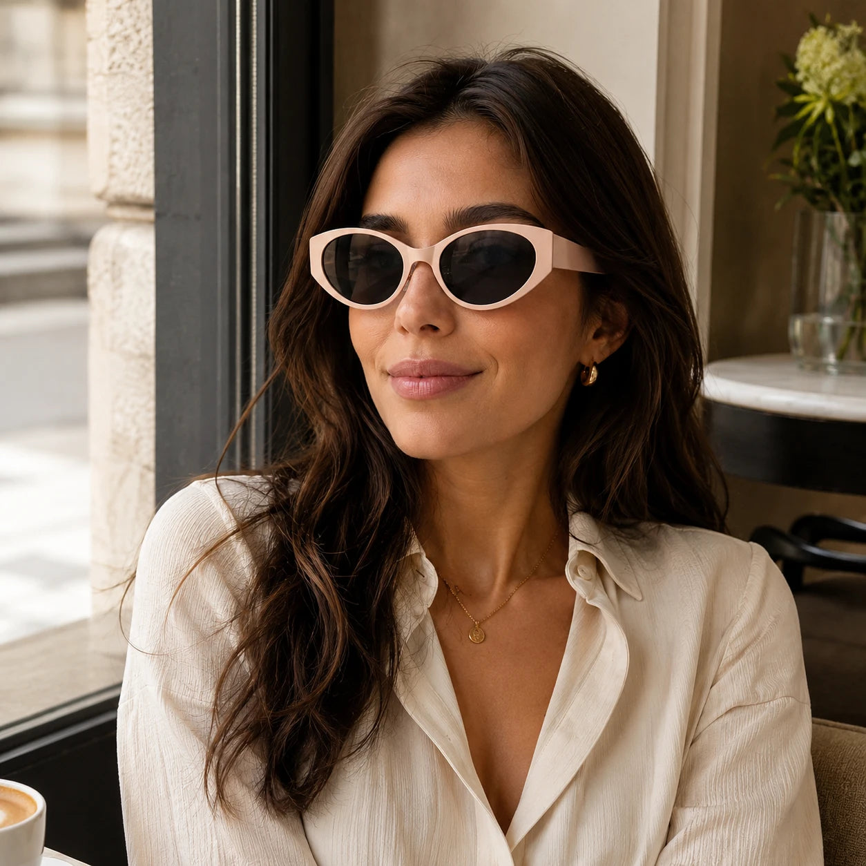 A chic woman wearing Lyra nude pink retro sunglasses while sitting by a window at a coffee shop.