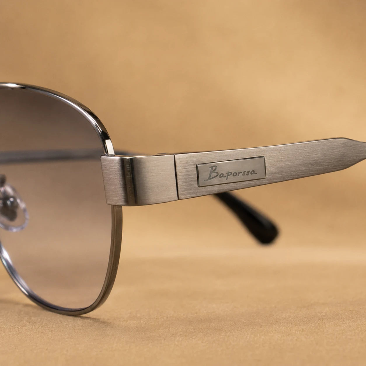 Close-up of BAPORSSA Aurelia gunmetal sunglasses showing brushed metal temple, logo plate, hinge detail, and smoke lens edge