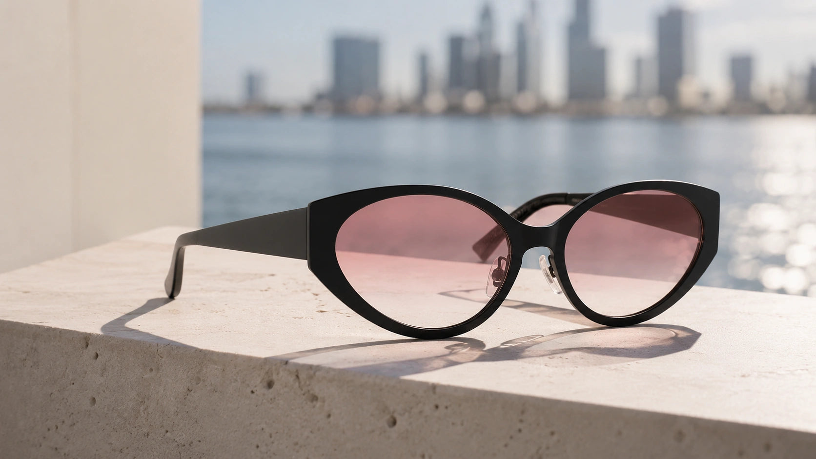 BAPORSSA Lyra black cat-eye sunglasses resting on a light stone
     surface with a blurred urban cityscape and waterfront in the background