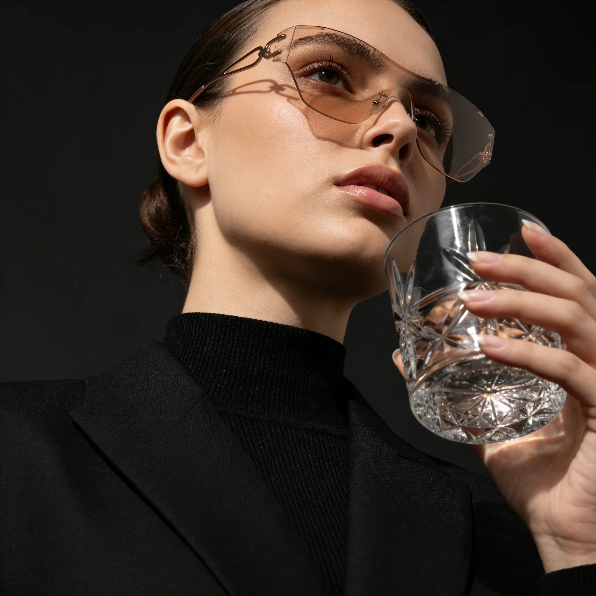 Person wearing sunglasses and holding a glass against a dark background