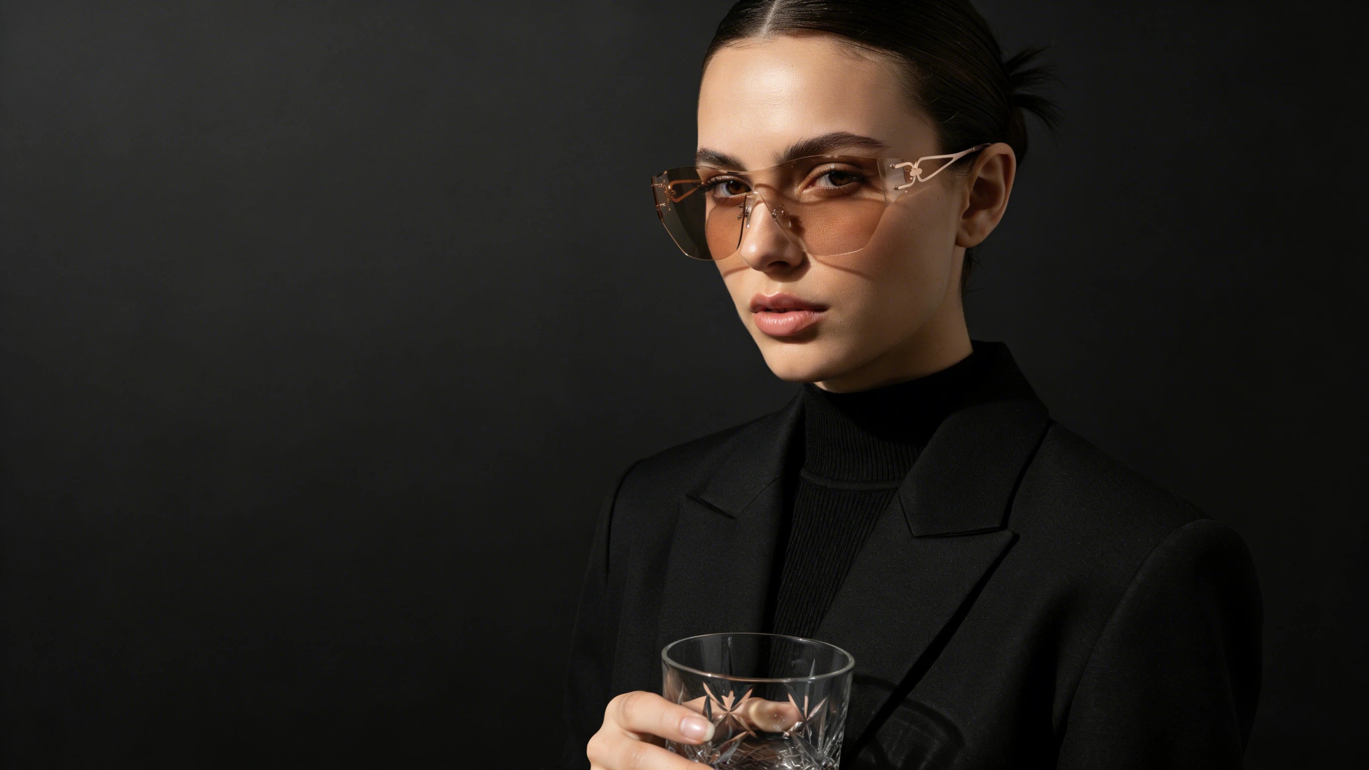 Woman wearing sunglasses and holding a glass against a black background