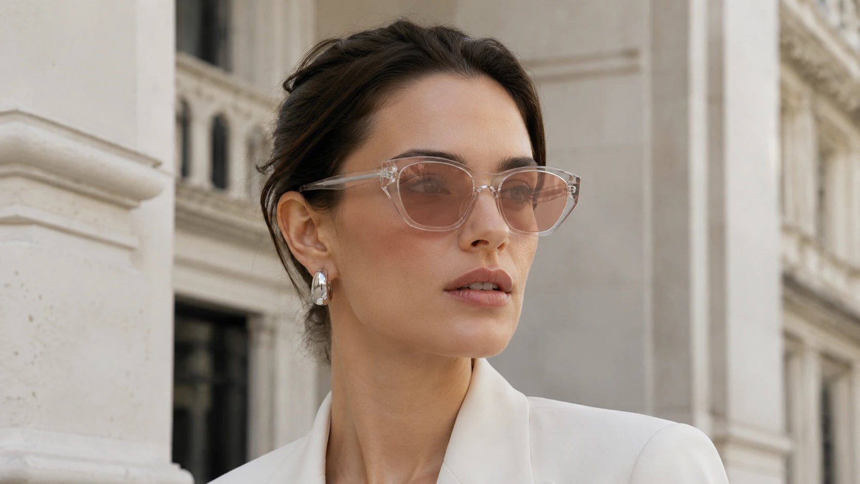 Woman wearing transparent Dune sculpted cat-eye sunglasses with soft rose nylon lenses, glossy acetate frame volume, and lifted outer corners in a refined city setting.