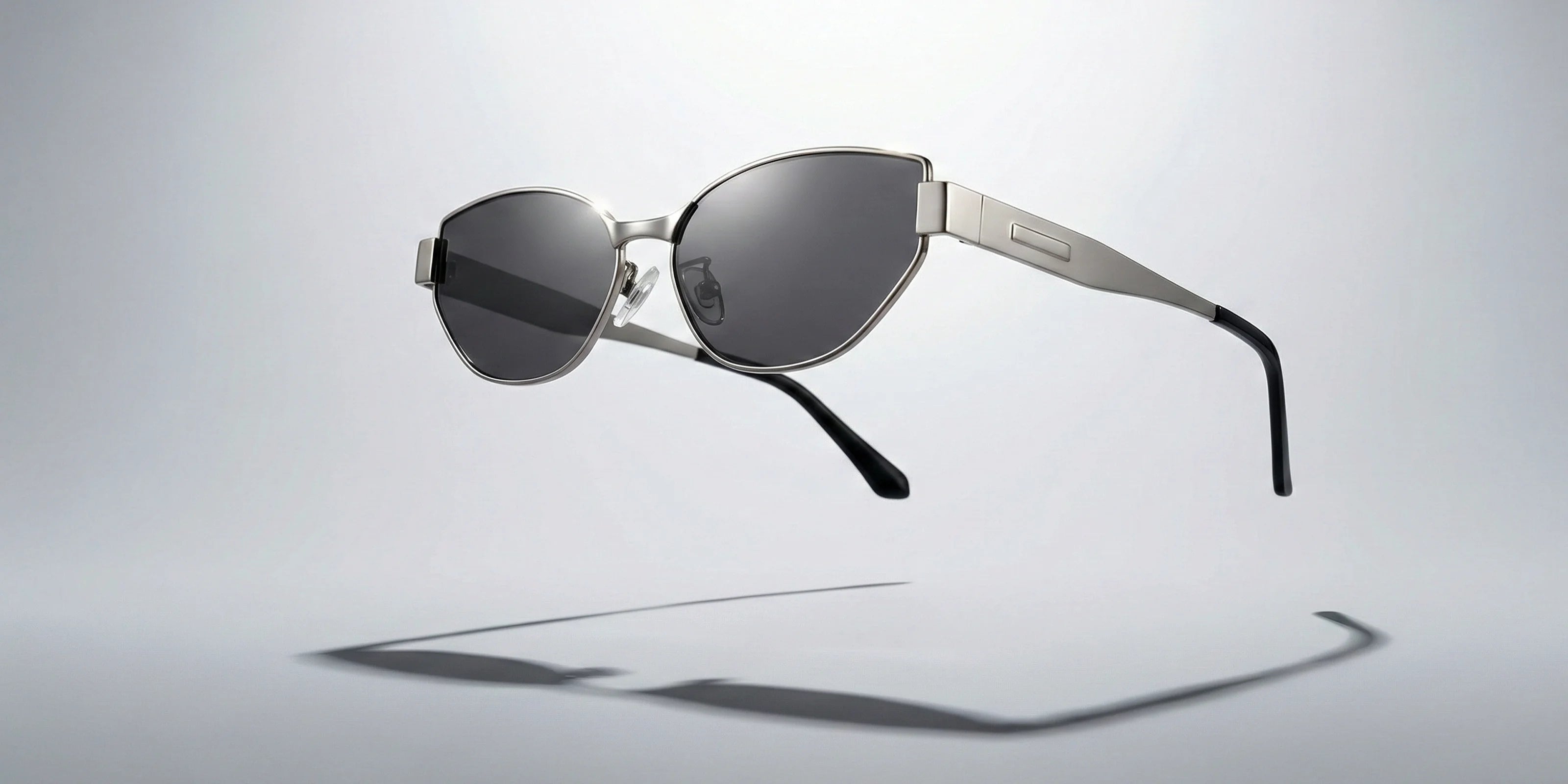 A pair of cat-eye sunglasses with a polished metal frame and dark lenses, floating against a minimalist grey background, showcasing high-end craftsmanship and avant-garde style.