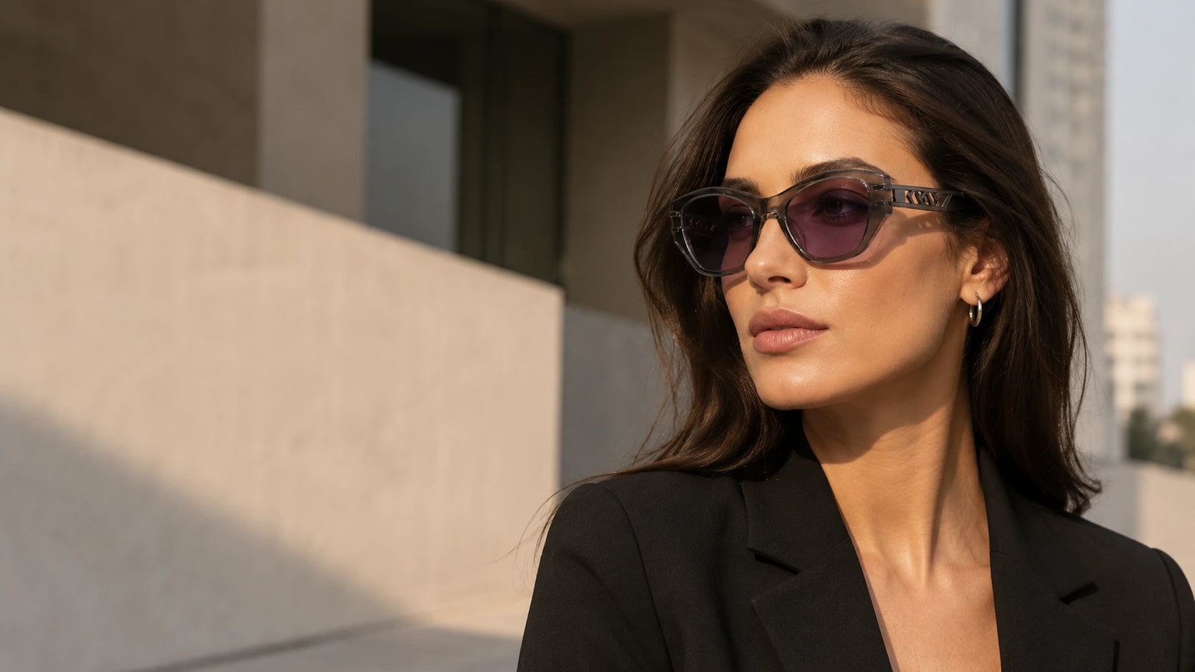 Woman wearing Noir structured cat-eye sunglasses with purple nylon lenses, translucent grey acetate frame, and cut-out metal temple details in warm city light.