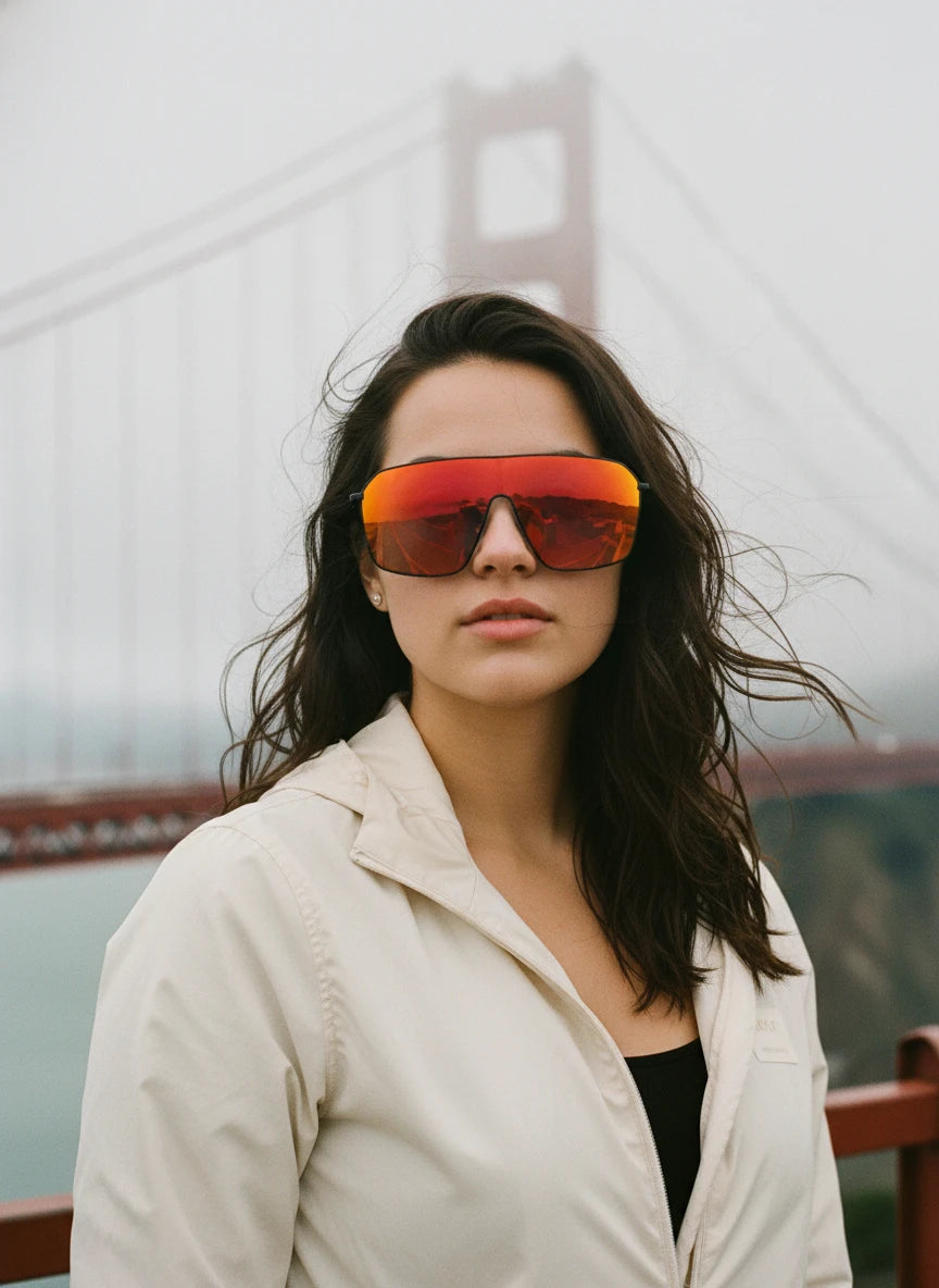 A woman wearing red mirror shield sunglasses riding in a convertible along a coastal highway, showcasing a bright summer travel eyewear style.