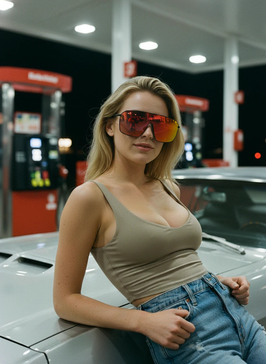 A woman wearing oversized red mirror shield sunglasses leaning against a car at a gas station at night, showcasing a bold urban fashion eyewear style.