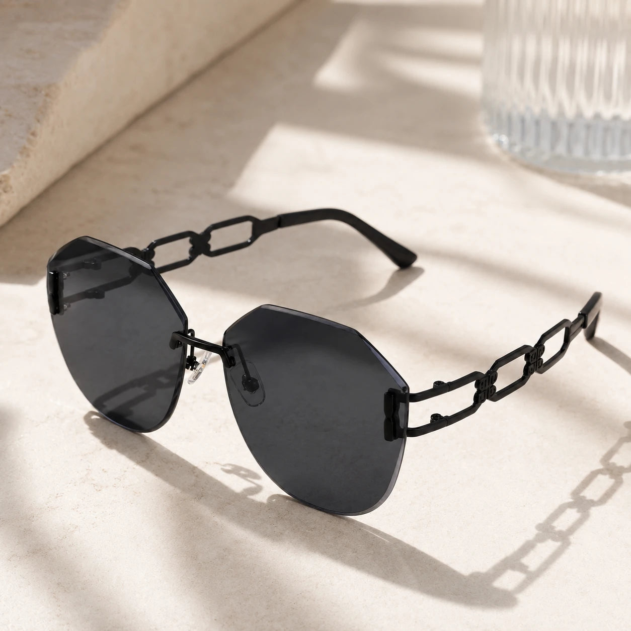 A pair of Baporssa arc oversized geometric polygonal rimless sunglasses with dark black lenses and intricate black hollowed geometric chain-link temples, resting on a sunlit stone window sill.