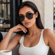 A portrait of a woman wearing Baporssa arc rimless black geometric sunglasses, sitting at a cafe table with her hand on her chin.