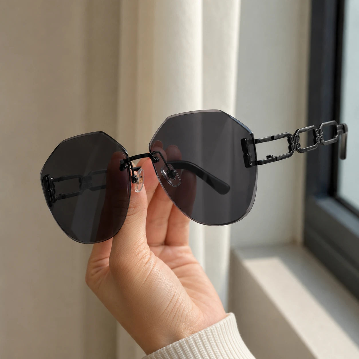 A close-up view of a hand holding Baporssa arc geometric rimless sunglasses, focusing on the detailed black hollowed geometric chain-link temple against a blurry background.