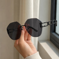 A close-up view of a hand holding Baporssa arc geometric rimless sunglasses, focusing on the detailed black hollowed geometric chain-link temple against a blurry background.