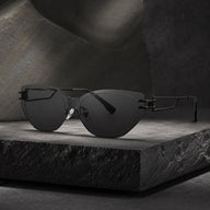 A premium still life shot of Baporssa Edge rimless cat-eye sunglasses with sharp dark lenses and geometric black metal temples, resting on a textured dark stone block.