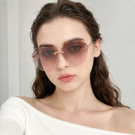 A direct front view portrait of a woman wearing Baporssa Glow rimless sunglasses with pinkish-brown gradient polygonal lenses and an off-the-shoulder white top.