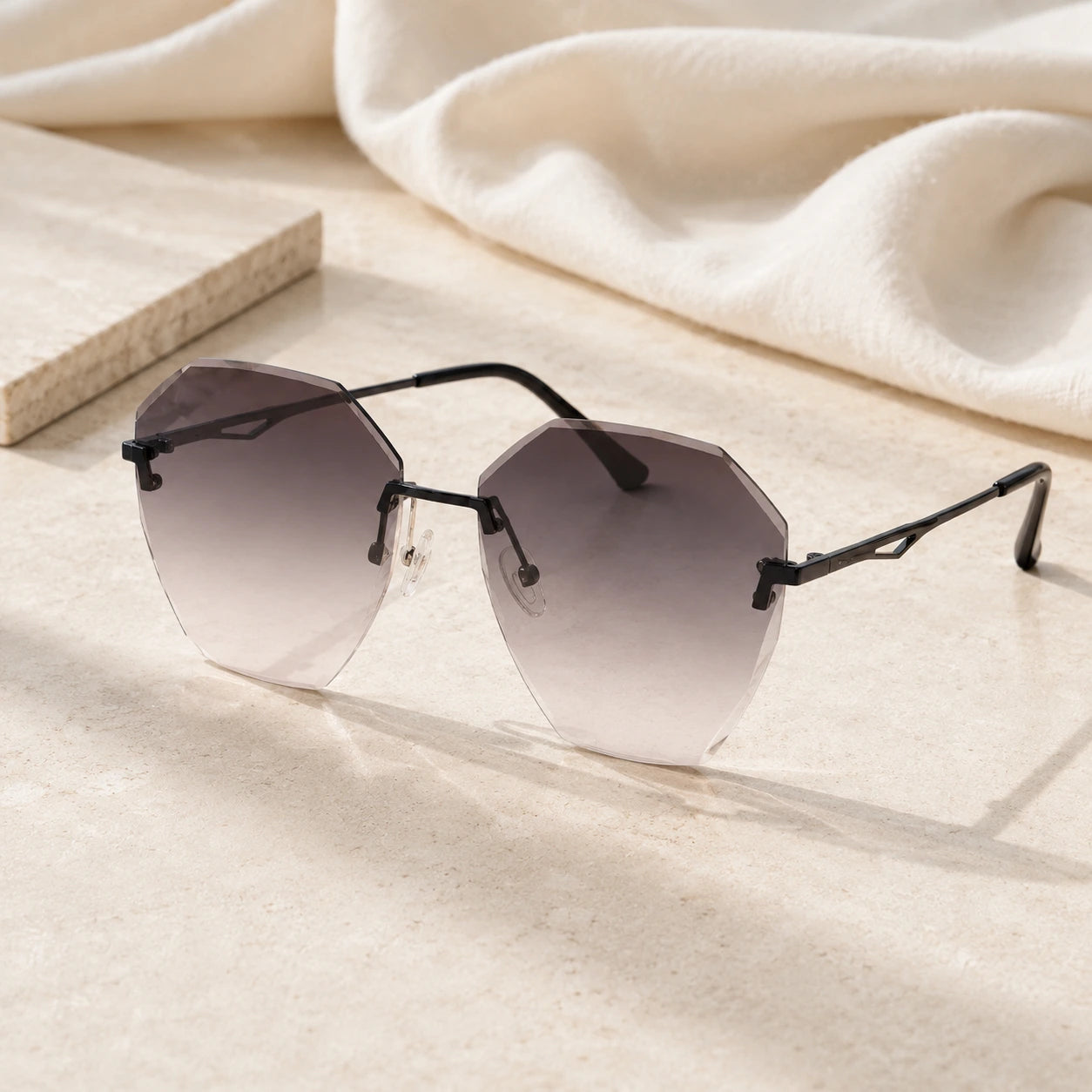 Baporssa Glow rimless polygonal sunglasses with dark grey gradient lenses and black hardware, displayed on a sunlit beige stone tile with a white linen background.