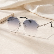 Baporssa Glow rimless polygonal sunglasses with blue-gray gradient lenses and silver hardware, resting on a sunlit marble surface next to a soft cream fabric.