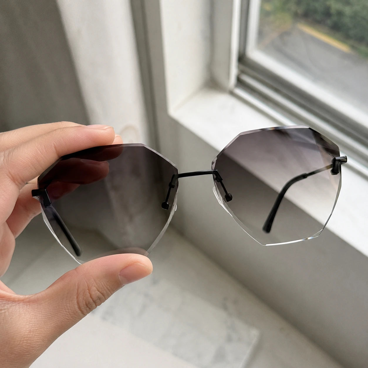 A close-up of a hand holding Baporssa Glow rimless geometric sunglasses against a bright window, showcasing the clarity of the gray gradient lenses.