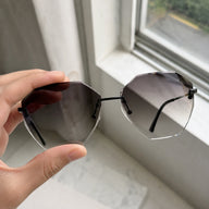 A close-up of a hand holding Baporssa Glow rimless geometric sunglasses against a bright window, showcasing the clarity of the gray gradient lenses.