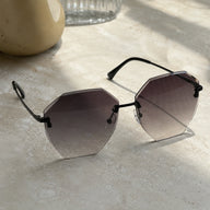 Baporssa Glow rimless polygonal sunglasses with dark gray gradient lenses and black metal hardware, resting on a sunlit beige marble counter with soft shadows.