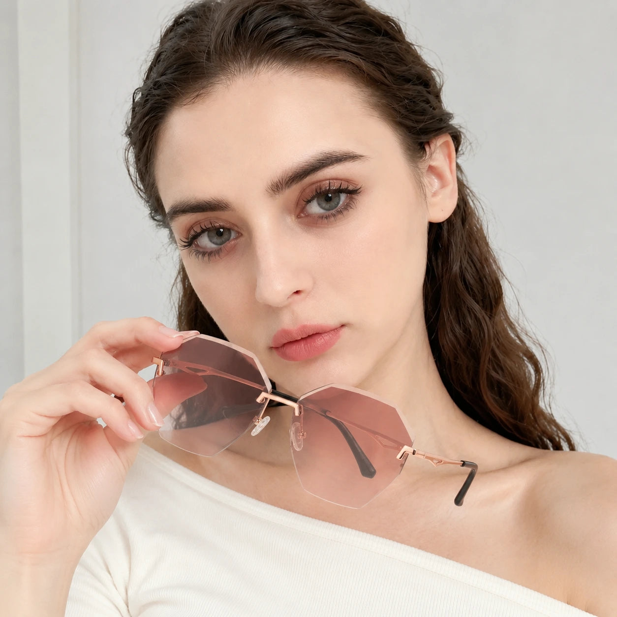 A close-up portrait of a woman with wavy hair holding Baporssa Glow rimless sunglasses with pinkish-brown gradient lenses and rose gold metal hardware.
