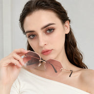 A close-up portrait of a woman with wavy hair holding Baporssa Glow rimless sunglasses with pinkish-brown gradient lenses and rose gold metal hardware.