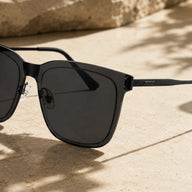A close-up side view of Baporssa Halo square sunglasses with a classic black frame and dark solid lenses, sitting on a sandy stone surface with shadows.