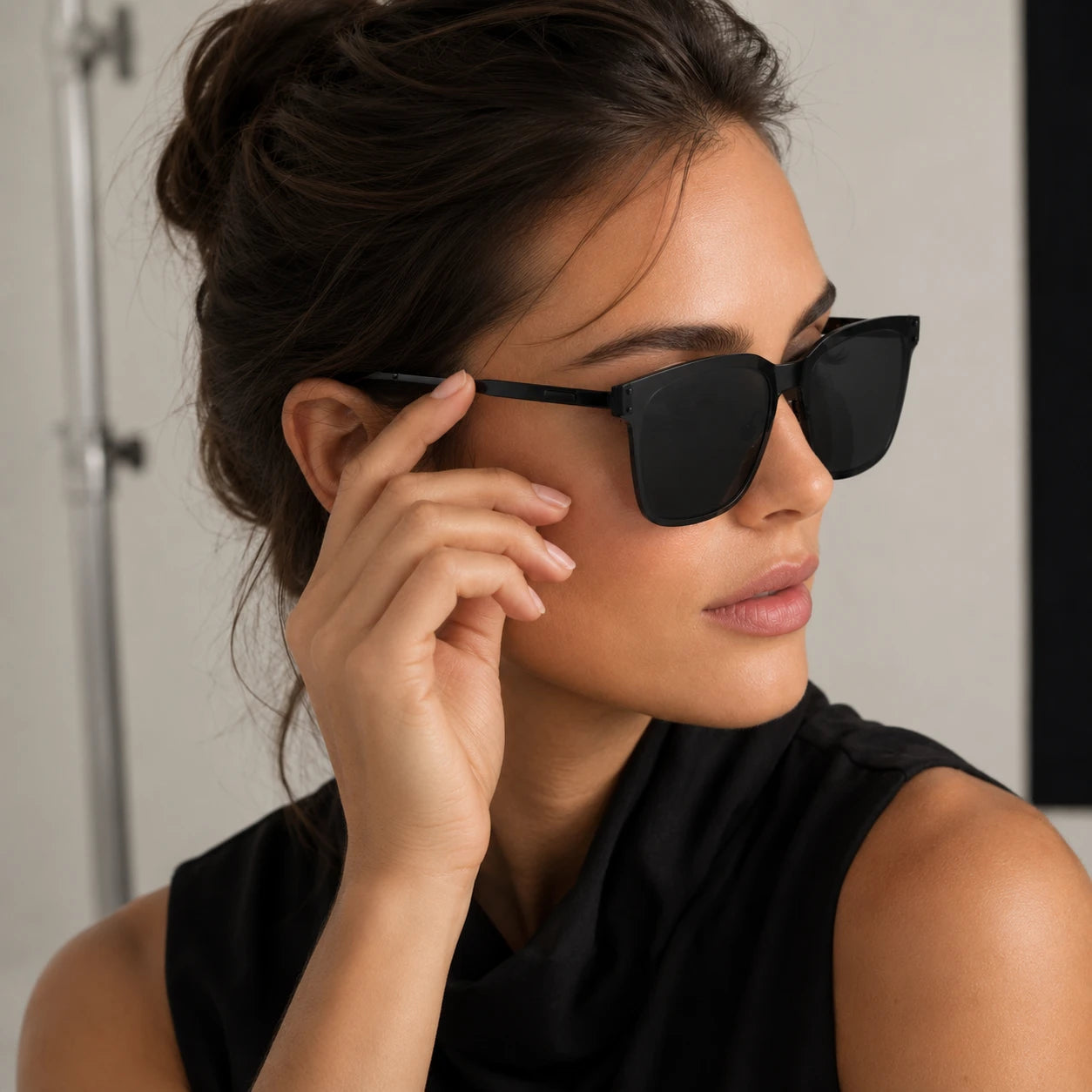 A studio portrait of a woman wearing Baporssa Halo black oversized sunglasses, with her hand gently resting near her temple arm.