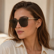 Three-quarter portrait of a woman looking away, wearing Baporssa Halo oversized square sunglasses with transparent jelly grey frames and gold temples.