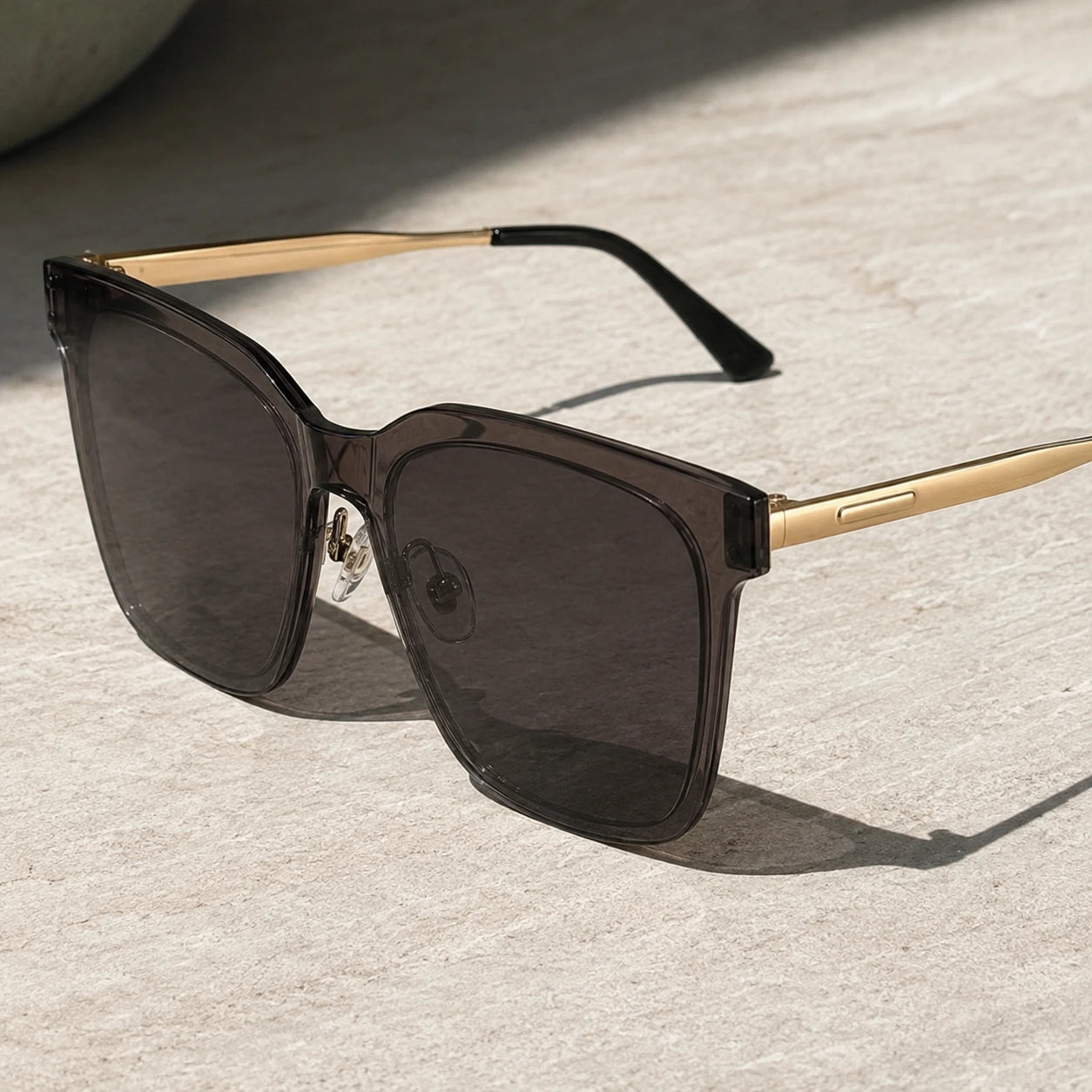 Angled product shot of Baporssa Halo sunglasses on a textured stone surface, highlighting the clear nose pads and the glossy finish of the grey frame.