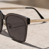 A tight close-up shot focusing on the hinge area of Baporssa Halo square sunglasses, highlighting the translucent grey acetate frame, dark lens, gold metal temple, and the metal nose pad hardware.