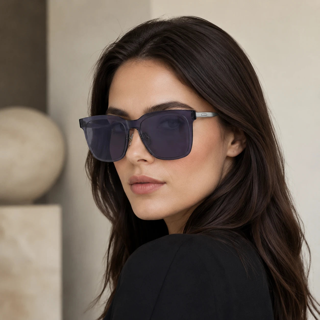 A close-up profile portrait of a woman looking over her shoulder, wearing Baporssa Halo square sunglasses with translucent dark grey frames and silver metal temples.