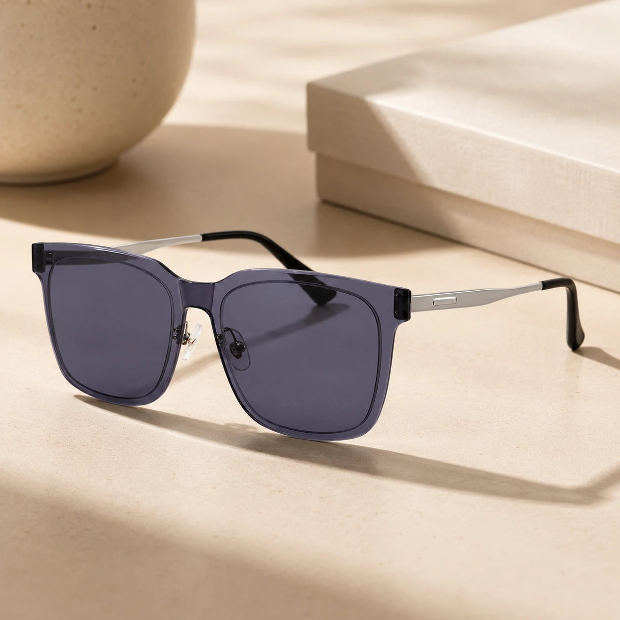A still life shot of Baporssa Halo square sunglasses with translucent dark grey frames and silver temples, placed on a beige surface with soft shadows next to a gift box.