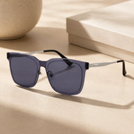 A still life shot of Baporssa Halo square sunglasses with translucent dark grey frames and silver temples, placed on a beige surface with soft shadows next to a gift box.
