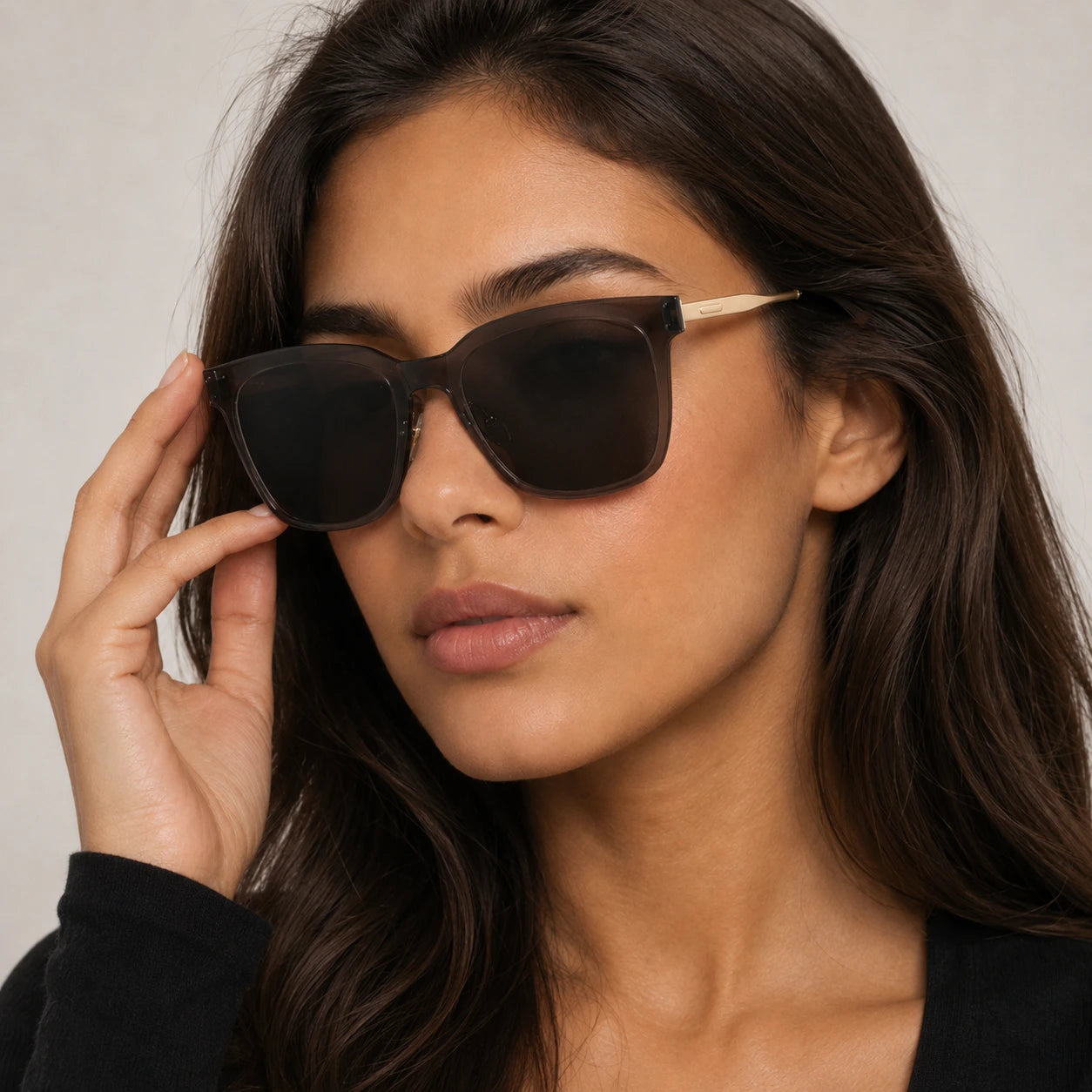 A close-up portrait of a woman wearing Baporssa Halo square sunglasses, gently touching the translucent grey frame.