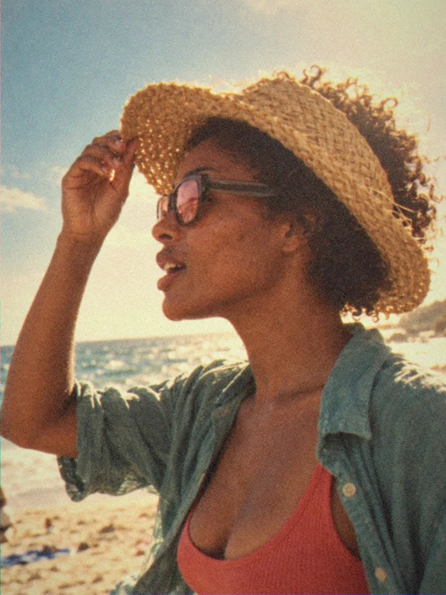 A woman in BAPORSSA Dune sunglasses with rose lenses holding her straw hat on a windy beach