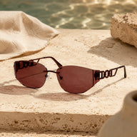 High-end product photography of Baporssa Muse rimless sunglasses with dark plum lenses, resting on a beige travertine stone next to rippling pool water.