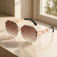 A pair of Baporssa rimless oversized sunglasses with brown gradient lenses and rose gold hollow geometric chain temples, resting on a sunlit stone window sill.