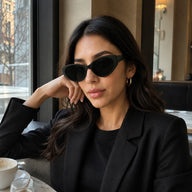 A chic woman wearing Lyra oversized black cat-eye sunglasses sitting by a window in a cafe.